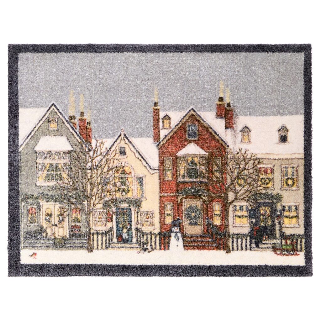 A rectangular doormat featuring a winter village scene with snow-covered houses, bare trees, storefronts, and a gray sky, finished with a dark border.
