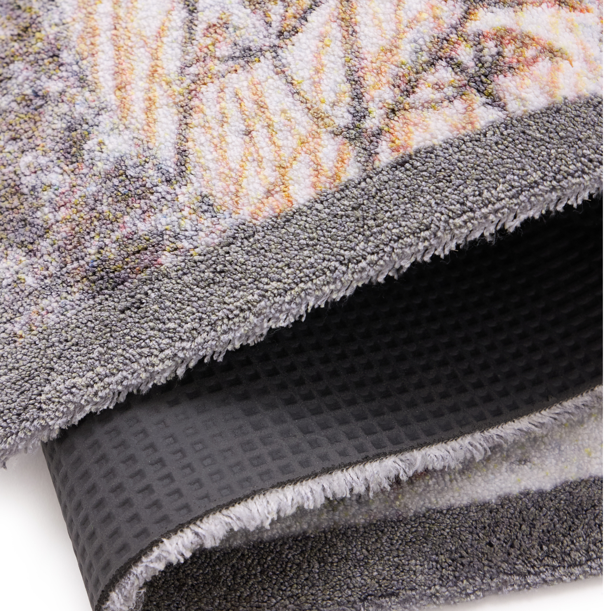 Close-up of a folded doormat showing a multicolor abstract pattern in gray, beige, and orange tones, with a dark gray textured backing visible underneath.