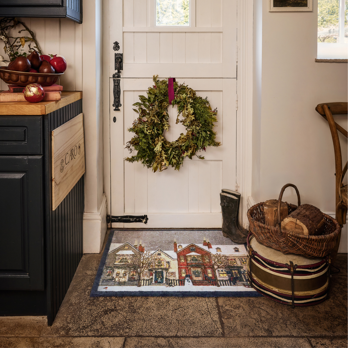 A holiday-themed doormat with a winter village scene, placed in front of a white front door in a rustic entryway. A green wreath hangs on the door, and a dark cabinet, basket, and wood stack are visible around it.