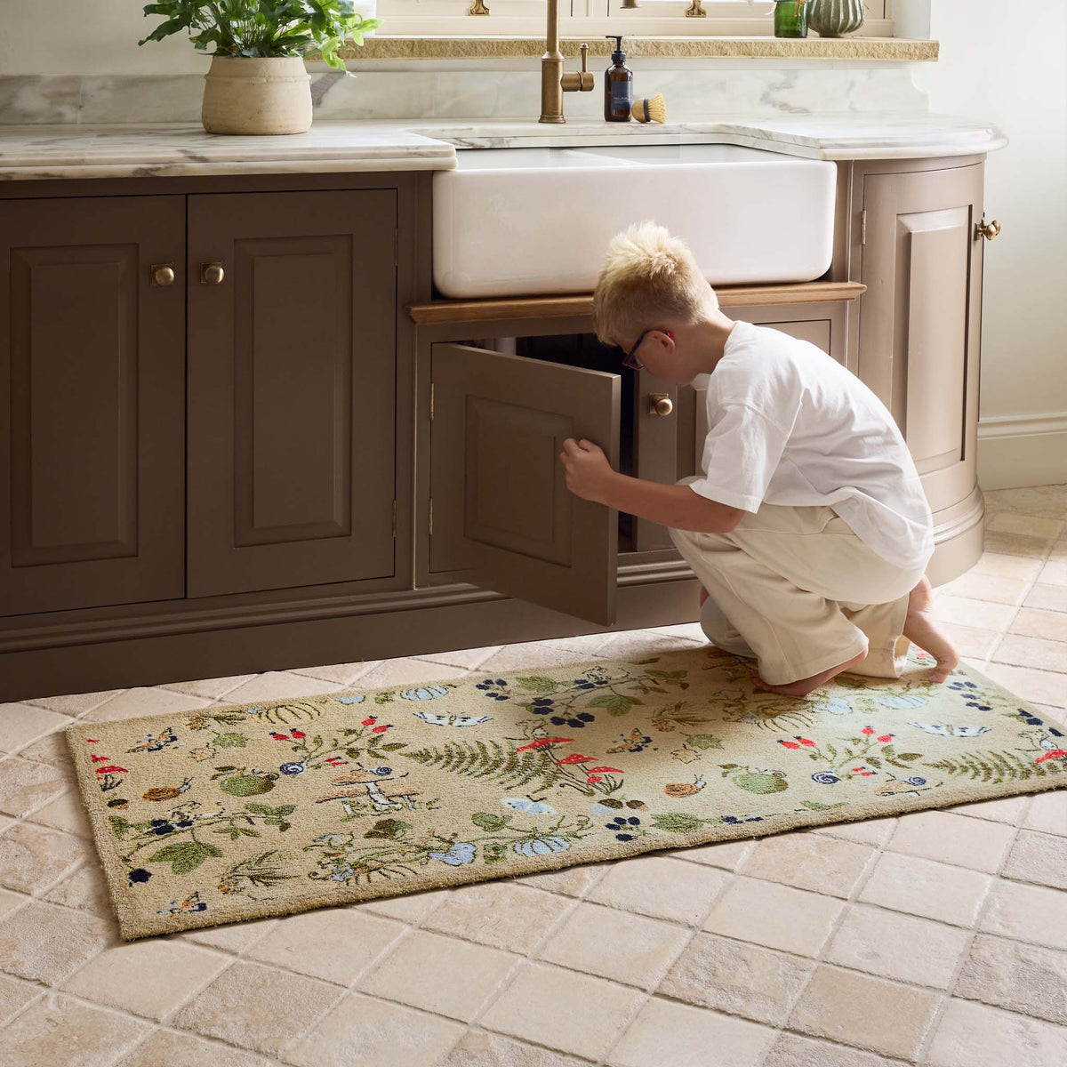A rectangular beige runner with a colorful  floral and botanical pattern, shown on a tiled kitchen floor in front of a brown vanity and white farmhouse sink.