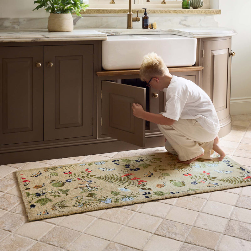 A rectangular beige runner with a colorful  floral and botanical pattern, shown on a tiled kitchen floor in front of a brown vanity and white farmhouse sink.