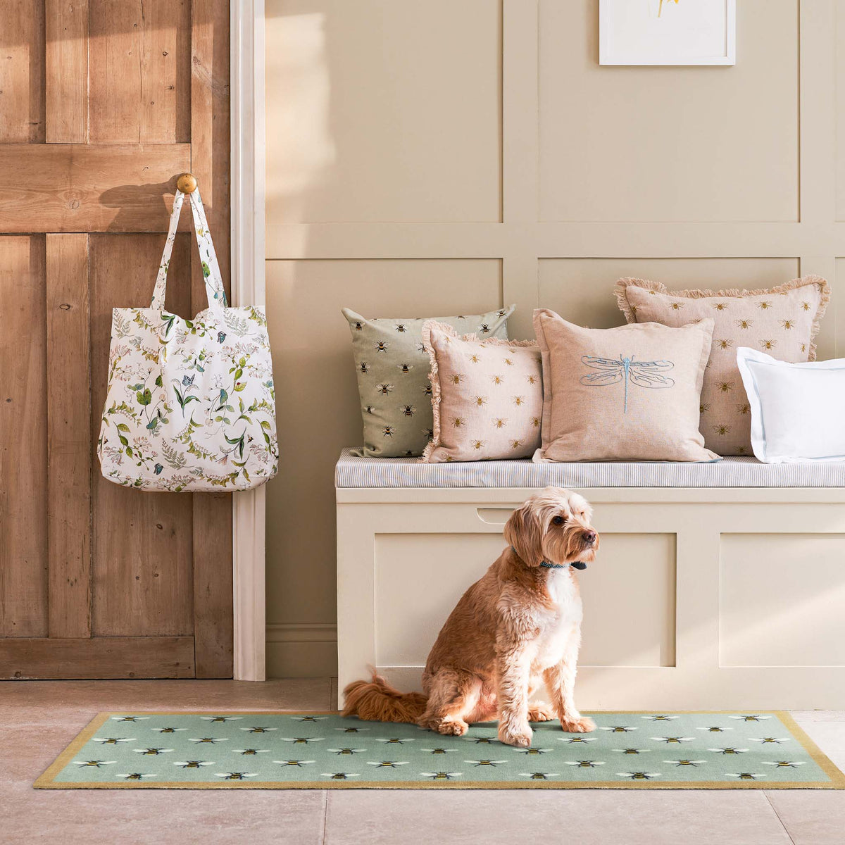 A dog sits on a blue patterned runner in a sunlit entryway, with a white tote bag hanging on a wooden door and decorative pillows on a bench behind it.