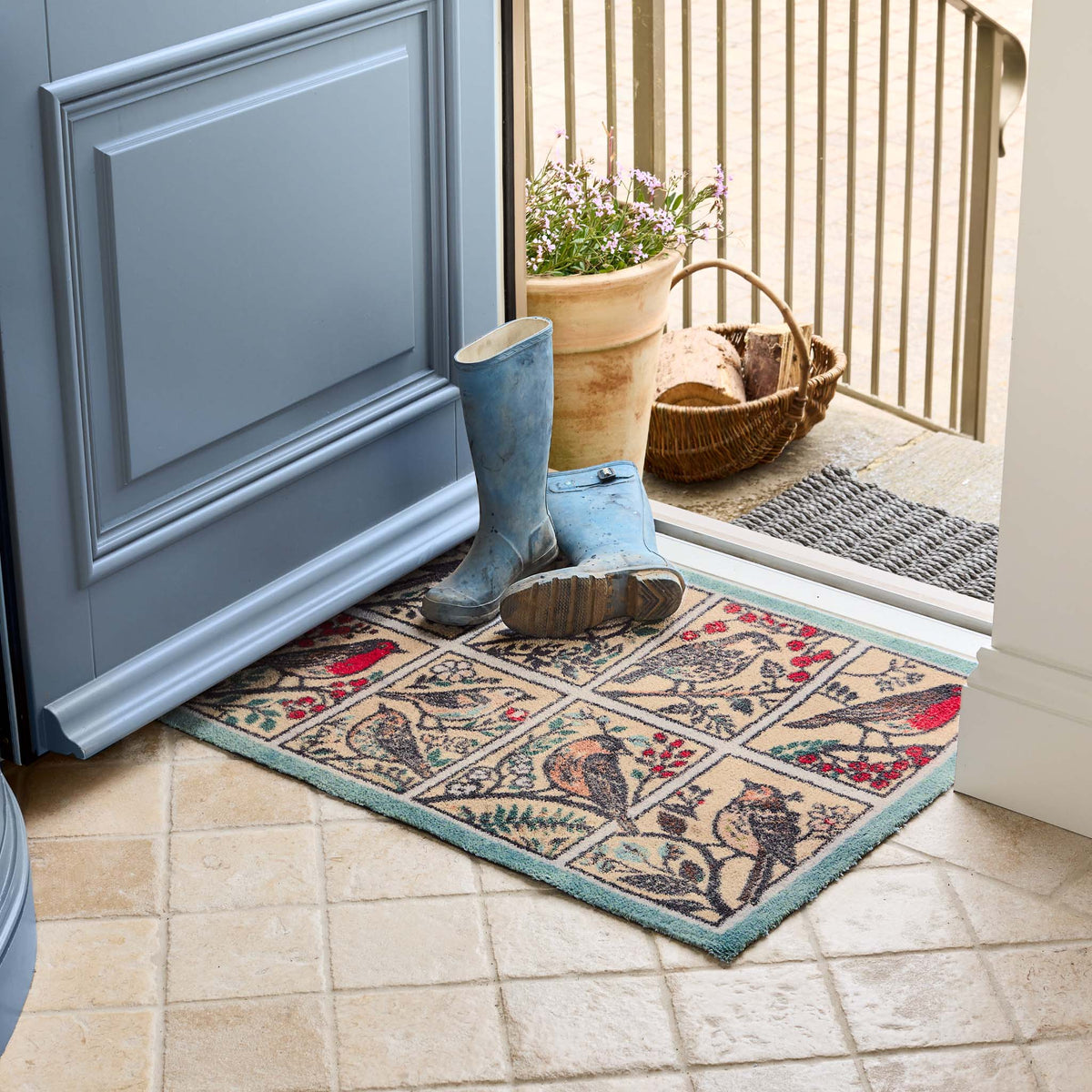 A rectangular doormat with a vintage-style bird panel design in beige, teal, red, and brown tones, placed in front of a blue door. A pair of blue rain boots and a wicker basket sit on the mat at the doorway.