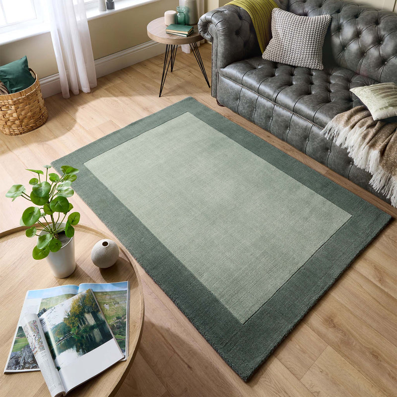 Borders Sage Green Rug - 100% wool rug in light green with a darker green border, styled in a living room. 