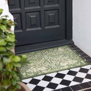 A rectangular outdoor doormat with an ornate green and beige damask pattern, placed on a tiled porch step in front of a black front door.