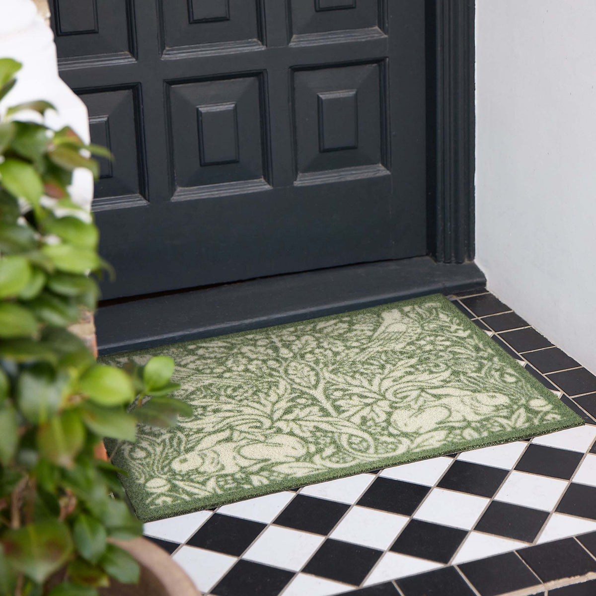 A rectangular outdoor doormat with an ornate green and beige damask pattern, placed on a tiled porch step in front of a black front door.