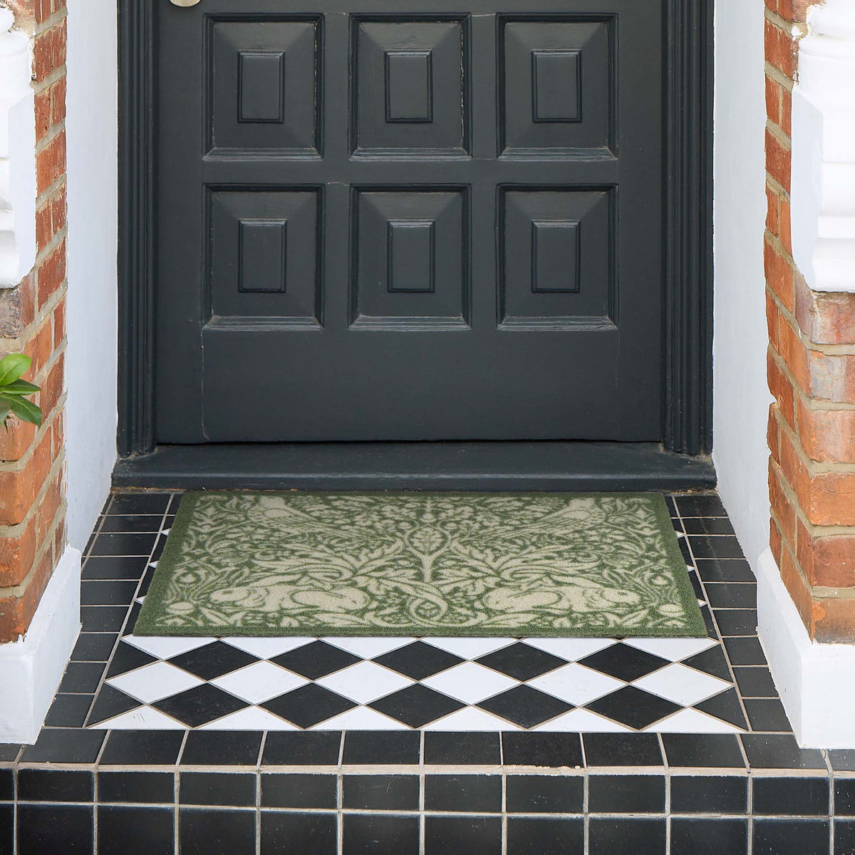 A rectangular outdoor doormat with an ornate green and beige damask pattern, placed on a tiled porch step in front of a black front door.