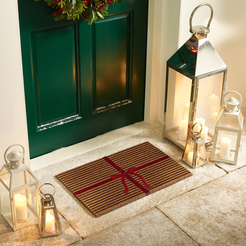 Discover the Christmas Gift Wrapped Coir Doormat- a coir doormat featuring a bright red bow and red lines giving the illusion of a present. 
