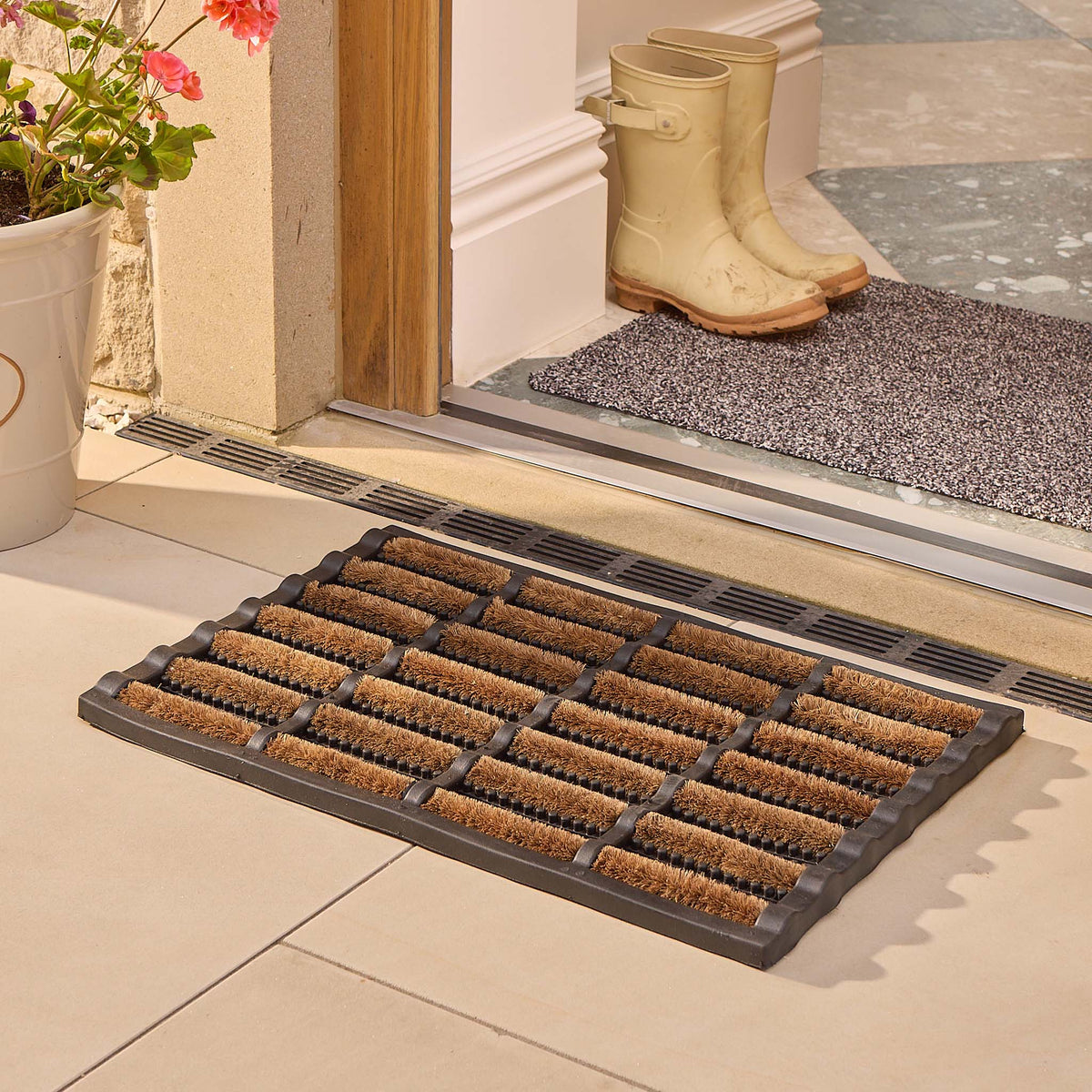A rectangular black rubber doormat with multiple angled brown bristles sits on a tiled entryway floor in front of a doorway. A second mat and a pair of cream rain boots are visible on the porch step, with a potted plant to the left.