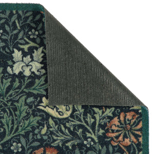 A close-up of a patterned doormat corner with the top-left corner folded back to show the dark gray backing. The rug features a dark navy blue background with a detailed botanical and floral pattern in green, cream, and rust orange.