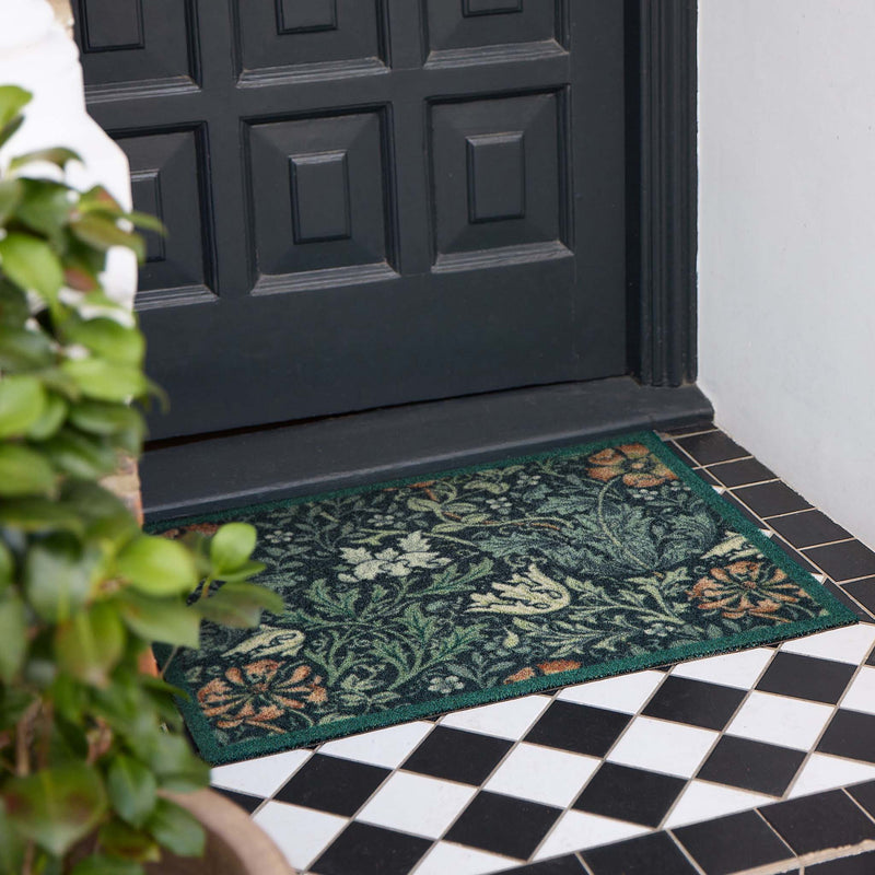 A rectangular floral outdoor doormat in dark green with orange and cream botanical patterns, placed on a tiled entryway in front of a black front door.