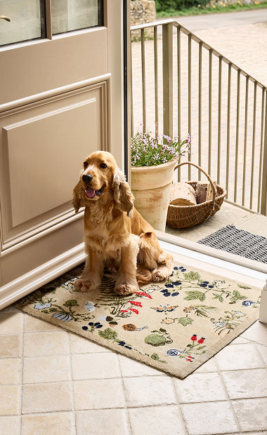 Doormat in an open doorway with a dog sitting on the mat. The mat has an Autumnal pattern on it, with a beige background. 