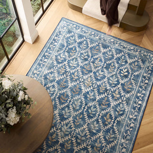 A rectangular blue area rug with a cream and beige botanical lattice pattern, shown on a light wood floor in a bright room with a window, table, and staircase visible.