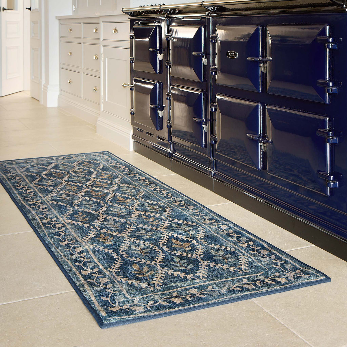A rectangular blue area rug with a traditional floral and geometric pattern, shown on a light beige tiled kitchen floor next to a glossy blue Aga stove.