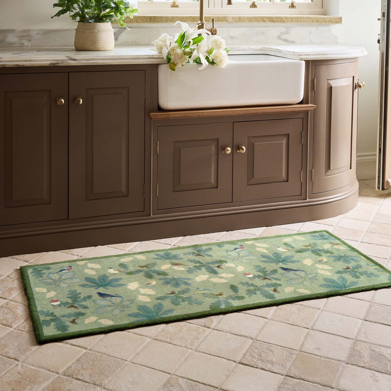 A green runner with a leaf and bird pattern lies on a beige tiled floor in front of a brown kitchen cabinet with a white farmhouse sink.