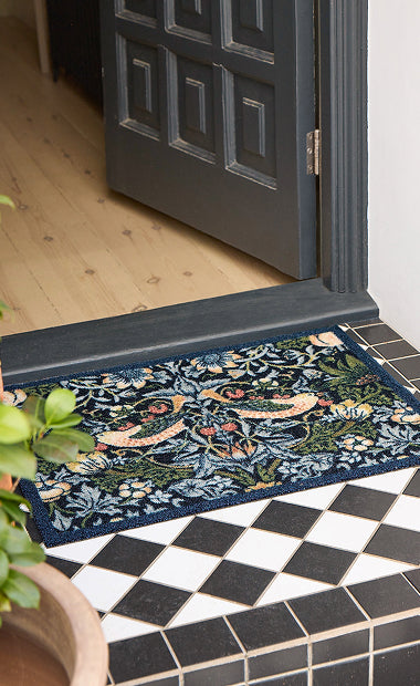 Morris & Co. outdoor mat, in the Strawberry Thief design with a navy border. On the floor outside an open door. 