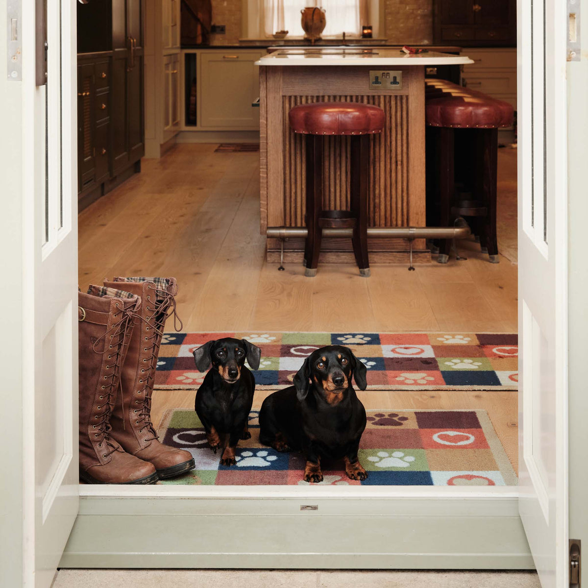 Two dachshunds sitting on a colorful paw-print patterned doormat in a kitchen doorway, with brown boots on the left and a kitchen island in the background.