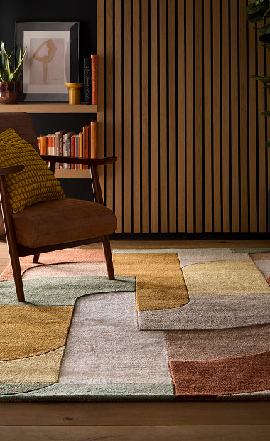 A modern and retro area rug with a geometric design in muted mustard,  green, taupe and cream, styled in a contemporary living room next to a brown armchair.