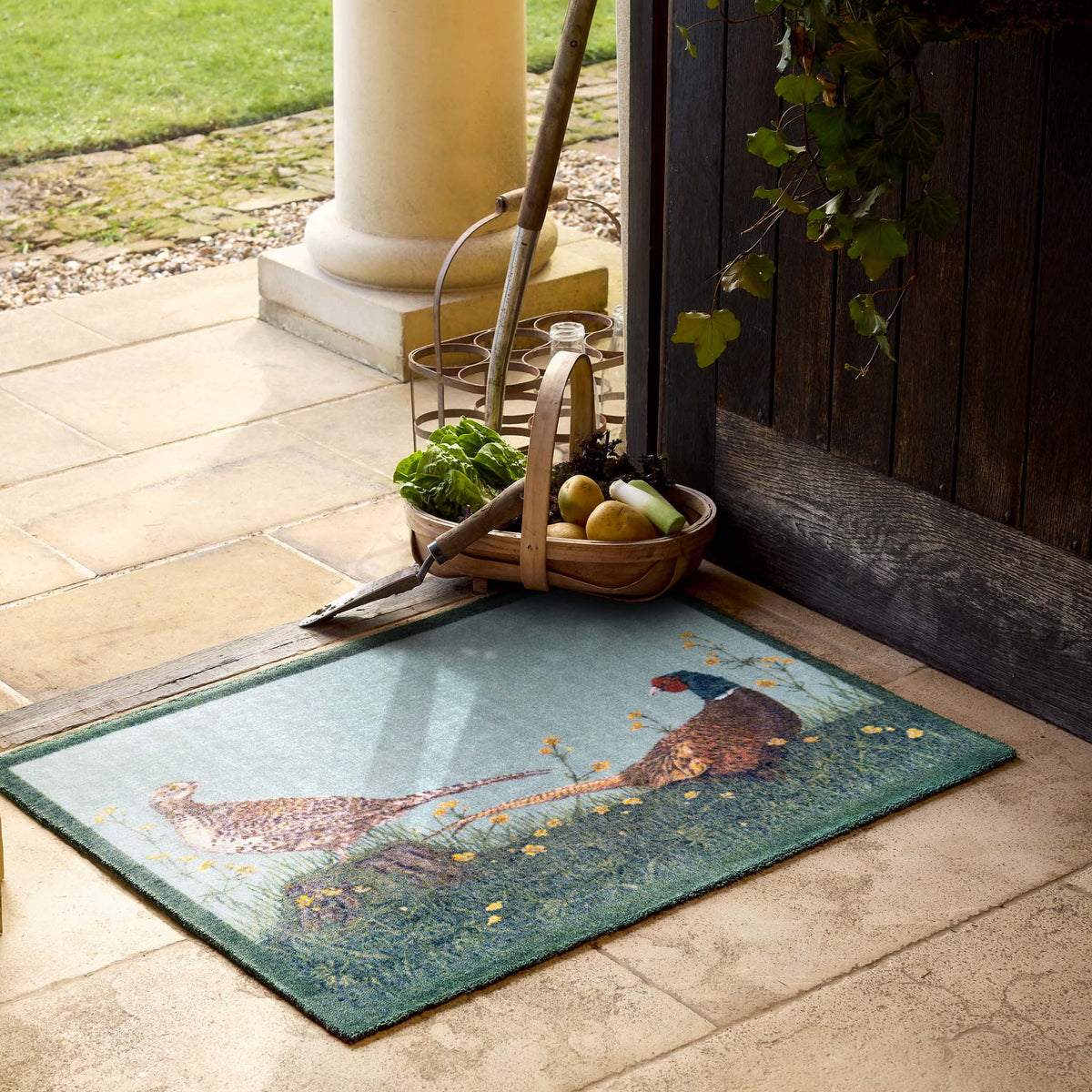 A rectangular doormat with a pheasant hunting scene, placed on a stone patio in front of a wooden door. A basket of produce, a garden tool, and a column are visible in the background.