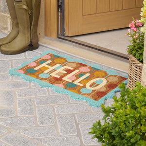 A colorful doormat with the word 'HELLO' in cream letters, surrounded by pastel fruit and a turquoise border, placed on a stone patio in front of a wooden door. Olive green rain boots and potted plants are visible nearby.