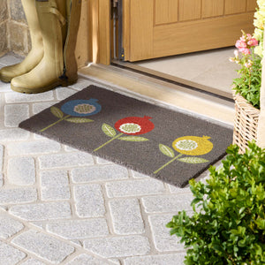 A rectangular dark grey doormat with a colorful flower pattern, placed at a wooden doorway on a stone patio. A pair of tall yellow rain boots stands on the left, and potted plants are visible on the right.
