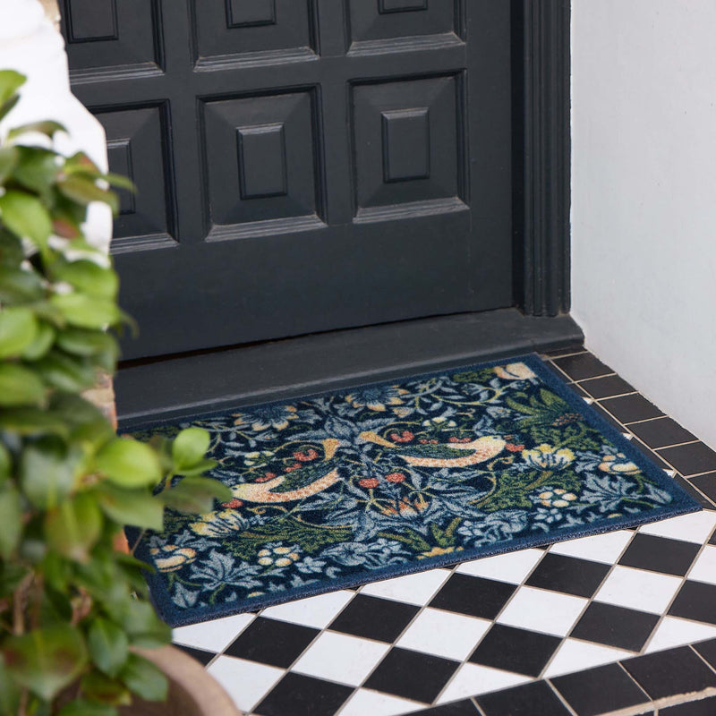 A rectangular outdoor doormat with an intricate floral and bird pattern in navy blue, green, beige, and orange, placed on a black and white tiled porch in front of a dark paneled door.