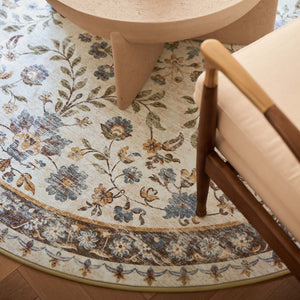 A round area rug with a vintage floral pattern in muted blue, beige, and brown tones, styled in a modern living room setting under a light wood table and next to a wooden armchair.