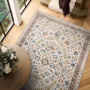 An area rug with a traditional floral pattern in blue, green, and gold on an ivory background, styled in a bright room with light wood floors, a round wooden table with flowers, and a staircase.