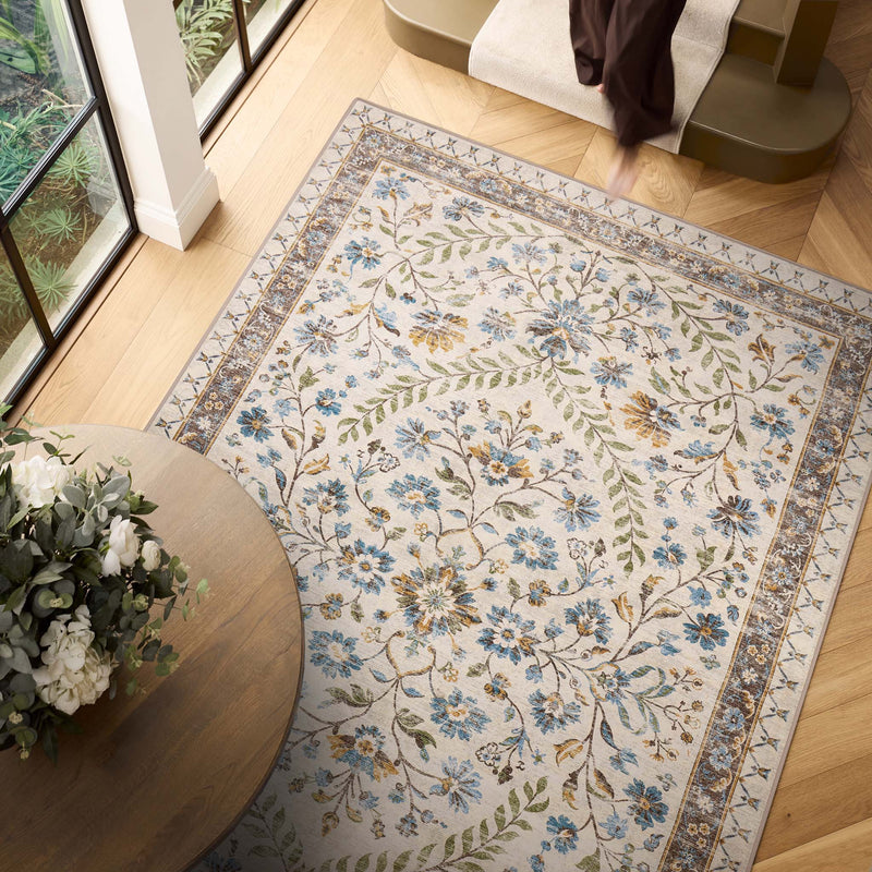 An area rug with a traditional floral pattern in blue, green, and gold on an ivory background, styled in a bright room with light wood floors, a round wooden table with flowers, and a staircase.