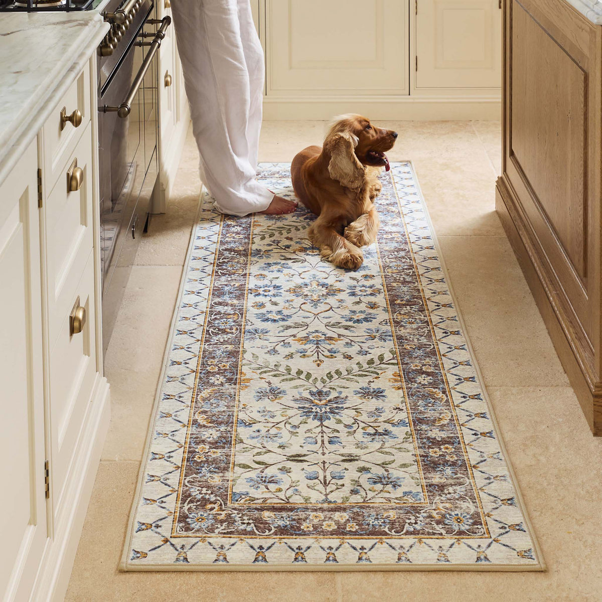 A long rectangular area rug with an ornate traditional floral pattern in cream, beige, muted blue, and olive green, shown flat against a solid white background.