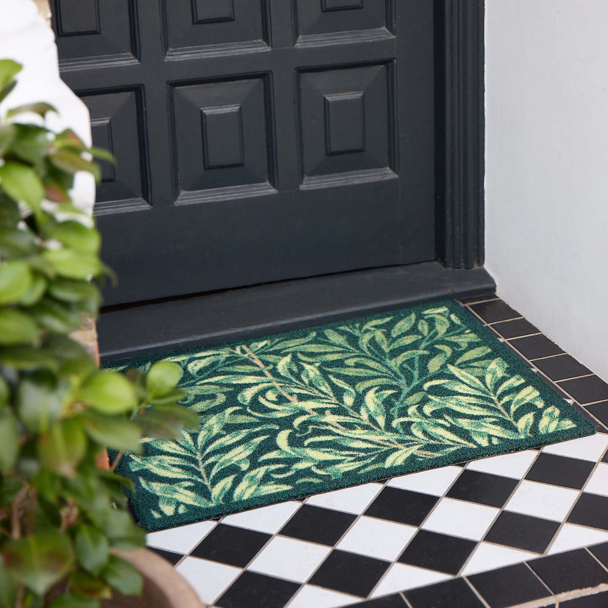 A rectangular green outdoor doormat with a leafy vine pattern, placed in front of a black paneled front door on a tiled porch.
